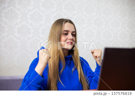 Happy blonde woman celebrates good news on laptop broadcast. She raises clenched fists in joy as positive report appears. Uplifting moment while watching television at home Happy blonde woman celebrates good news on laptop broadcast. She raises clenched fists in joy as positive report appears. Uplifting moment while watching television at home 137576819
