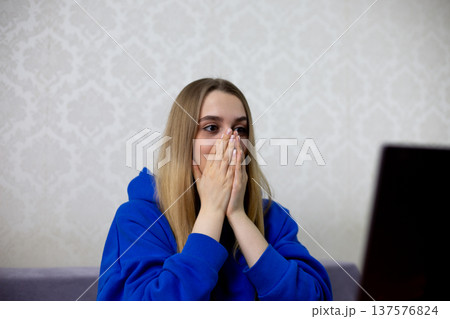 Shocked blonde football fan covers mouth and nose with both hands after incredible goal. She watches live match online, amazed by stunning play. Intense fan reaction at home 137576824