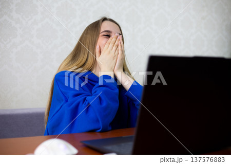 Young woman laughs joyfully during video call with friends on laptop. She covers face with both hands, sharing funny moment with loved ones online. Genuine connection and happiness at home 137576883