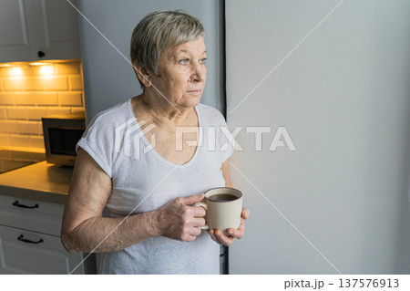 Senior woman looking out bright window, holding a coffee mug, thinking about life or future in a quiet moment 137576913