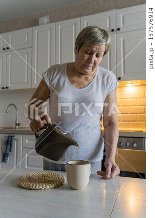 Senior woman in modern kitchen calmly pouring tea into a mug on the table, enjoying a peaceful morning routine and self-care. Real people, the life of an elderly person, daily habits 137576914