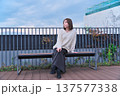 都会のテラスでベンチに座り微笑む女性モデル。冬の装いとリラックスした休日のひととき。 137577338