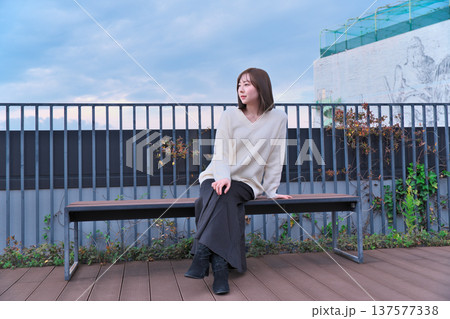 都会のテラスでベンチに座り微笑む女性モデル。冬の装いとリラックスした休日のひととき。 都会のテラスでベンチに座り微笑む女性モデル。冬の装いとリラックスした休日のひととき。 137577338