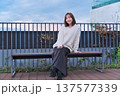 都会のテラスでベンチに座り微笑む女性モデル。冬の装いとリラックスした休日のひととき。 137577339