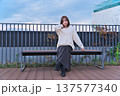 都会のテラスでベンチに座り微笑む女性モデル。冬の装いとリラックスした休日のひととき。 137577340
