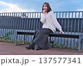 都会のテラスでベンチに座り微笑む女性モデル。冬の装いとリラックスした休日のひととき。 137577341