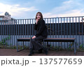 都会のテラスでベンチに座る黒コートの女性。洗練された冬の装いと穏やかな休日のポートレート。 137577659
