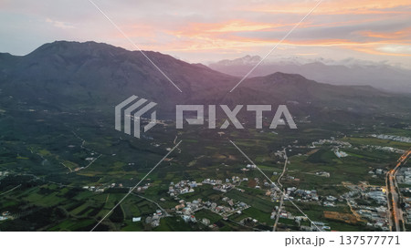 Aerial panoramic view of Crete foothills at sunset with snow-capped peaks, olive groves, valley with lake, scattered villages, winding road, and layered clouds Aerial panoramic view of Crete foothills at sunset with snow-capped peaks, olive groves, valley with lake, scattered villages, winding road, and layered clouds 137577771