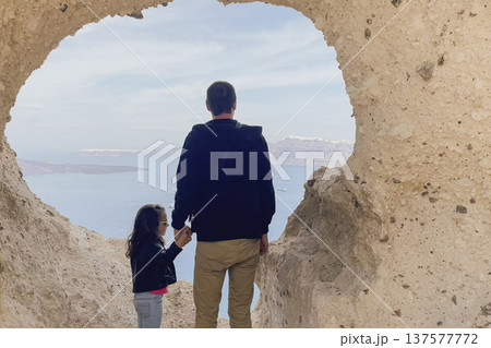 Father and young daughter holding hands, looking at Aegean Sea through heart-shaped natural rock arch on Santorini, with caldera and white houses in distance Father and young daughter holding hands, looking at Aegean Sea through heart-shaped natural rock arch on Santorini, with caldera and white houses in distance 137577772