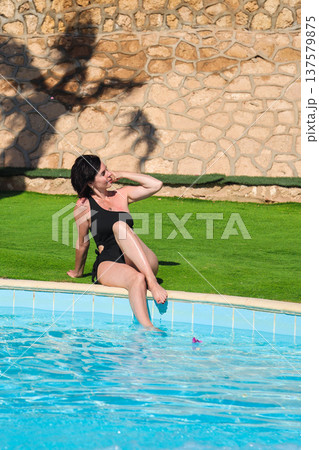 A woman in a bathing suit sits on the grass near a swimming pool with blue tiles A woman in a bathing suit sits on the grass near a swimming pool with blue tiles 137579875