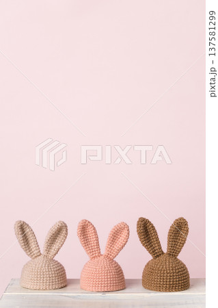 Three knitted bunny hats in pastel colors against a pink background Three knitted bunny hats in pastel colors against a pink background 137581299