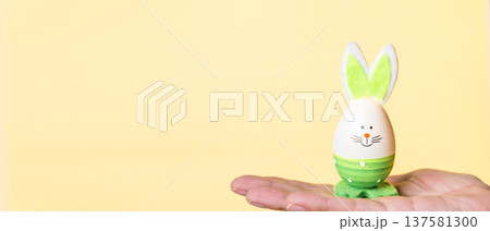 Decorative Easter egg with bunny ears held in hand against yellow background Decorative Easter egg with bunny ears held in hand against yellow background 137581300