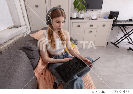Young woman using laptop on couch while wearing headphones in modern living room during daytime Young woman using laptop on couch while wearing headphones in modern living room during daytime 137581478