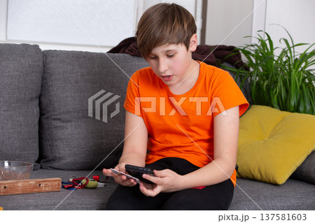Boy in orange shirt using smartphone while sitting on grey sofa at home Boy in orange shirt using smartphone while sitting on grey sofa at home 137581603