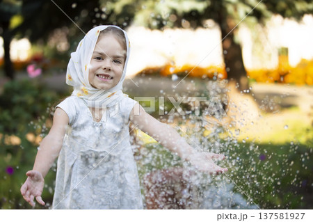 Young girl enjoys splashing water in a garden on a sunny day while wearing a light scarf Young girl enjoys splashing water in a garden on a sunny day while wearing a light scarf 137581927