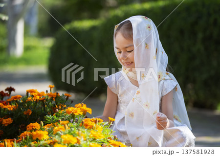 Young girl in a white scarf smiles while tending to orange marigold flowers in a sunny garden in spring Young girl in a white scarf smiles while tending to orange marigold flowers in a sunny garden in spring 137581928