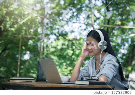 Asian woman wearing headphones listening to music on her laptop. Relaxing time. Lazy worker wasting time at workplace in co-working. Asian woman wearing headphones listening to music on her laptop. Relaxing time. Lazy worker wasting time at workplace in co-working. 137582903