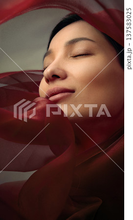 Close beauty portrait of Asian woman with eyes closed wrapped in flowing red silk fabric Close beauty portrait of Asian woman with eyes closed wrapped in flowing red silk fabric 137583025
