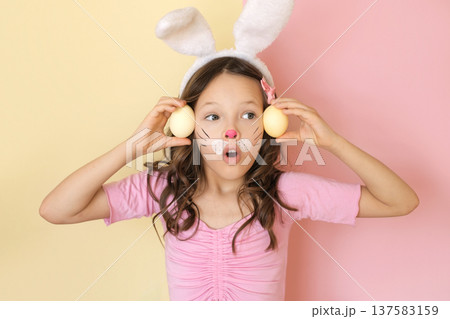 A girl with mouth open in surprise, shows yellow Easter eggs with a painted bunny face. A girl with mouth open in surprise, shows yellow Easter eggs with a painted bunny face. 137583159