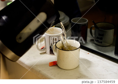 A man pours tea into two mugs on the kitchen table. The concept of comfort, hospitality, and everyday life, morning rituals, warmth, relaxation, and a homey lifestyle. A man pours tea into two mugs on the kitchen table. The concept of comfort, hospitality, and everyday life, morning rituals, warmth, relaxation, and a homey lifestyle. 137584459
