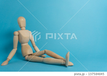 Wooden figure sits against a blue background with legs extended and body tilted slightly to one side during daylight hours Wooden figure sits against a blue background with legs extended and body tilted slightly to one side during daylight hours 137584845