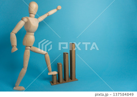 Wooden figure stands on one leg and raises arm next to cardboard bars against a blue background in a creative display of movement and growth 137584849