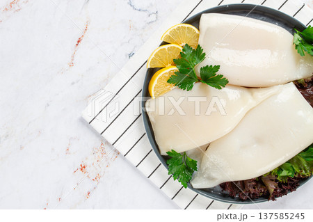 Fresh squid on a black plate with greens and lemon slices prepared for cooking in a kitchen setting Fresh squid on a black plate with greens and lemon slices prepared for cooking in a kitchen setting 137585245