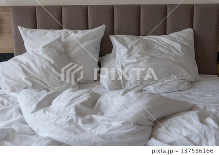 Unmade hotel bed with white pillows and rumpled duvet on mattress in modern bedroom interior. Hospitality concepts, hotel accommodation marketing, travel stay comfort, tourism lodging promotion 137586166