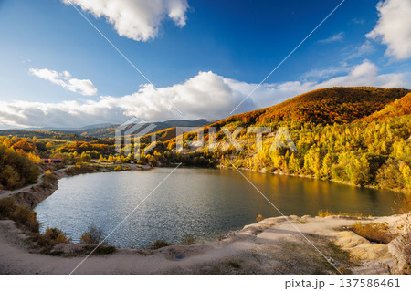 A lake nestled between autumn hills, with golden trees reflecting in the water under a bright blue sky and scattered clouds. Flooded quarry near the village Sutovo in Slovakia, Europe. A lake nestled between autumn hills, with golden trees reflecting in the water under a bright blue sky and scattered clouds. Flooded quarry near the village Sutovo in Slovakia, Europe. 137586461