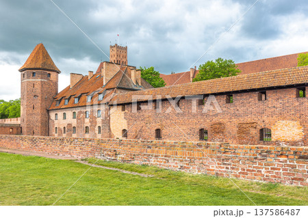 The Malbork Castle, a brick Gothic castle in northern Poland, Europe. Majestic medieval castle with red brick walls, towers, and tiled roofs, standing tall against a cloudy sky in a historic landscape The Malbork Castle, a brick Gothic castle in northern Poland, Europe. Majestic medieval castle with red brick walls, towers, and tiled roofs, standing tall against a cloudy sky in a historic landscape 137586487