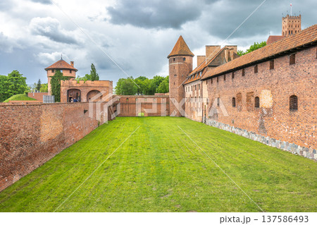 The Malbork Castle, a brick Gothic castle in northern Poland, Europe. Courtyard View of Historic Fortress: Brick walls, lush grass, towers under cloudy skies create an imposing architectural scene. 137586493