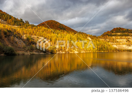 A serene lake reflects the golden hues of autumn foliage on the surrounding hills, casting a warm glow under a cloudy sky. Flooded quarry near the village Sutovo in Slovakia, Europe. A serene lake reflects the golden hues of autumn foliage on the surrounding hills, casting a warm glow under a cloudy sky. Flooded quarry near the village Sutovo in Slovakia, Europe. 137586496