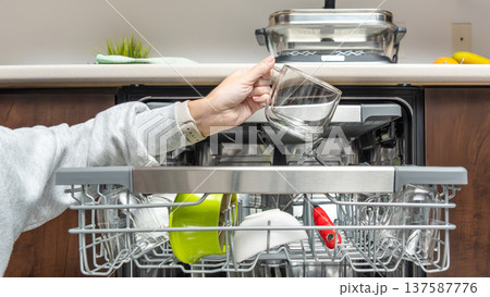 Hand placing glass cup into modern dishwasher filled with clean dishes in a contemporary kitchen setting with wooden cabinetry Hand placing glass cup into modern dishwasher filled with clean dishes in a contemporary kitchen setting with wooden cabinetry 137587776
