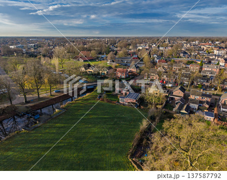 Panoramic aerial view of a charming European town with brick houses, a winding canal, and green meadows. A peaceful suburban landscape under a blue sky with soft clouds during the morning. Panoramic aerial view of a charming European town with brick houses, a winding canal, and green meadows. A peaceful suburban landscape under a blue sky with soft clouds during the morning. 137587792