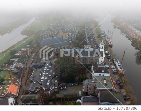 Aerial view of the historic Grote Kerk church in Muiden, Netherlands, surrounded by traditional houses and a canal shrouded in thick morning fog. A moody, atmospheric Dutch cityscape from above. 137587806