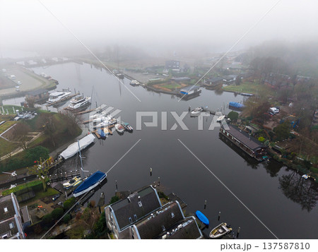 Aerial view of a foggy harbor in Muiden, Netherlands. Modern yachts and houseboats line the misty canal in a calm, atmospheric Dutch morning landscape. High angle drone perspective. 137587810