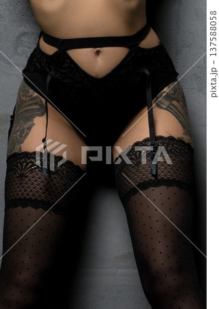 Model poses in black lingerie with straps and thigh-high stockings against a gray wall in an indoor setting 137588058