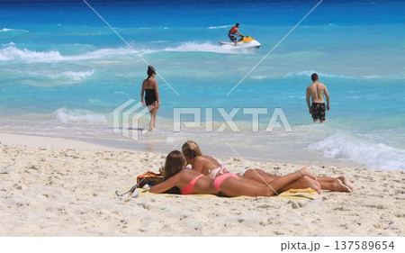 Two women sunbathe on sandy beach while other visitors wade in clear turquoise water Two women sunbathe on sandy beach while other visitors wade in clear turquoise water 137589654