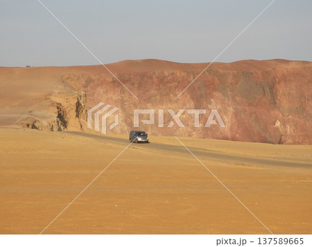 Silver van driving along a narrow paved road through a vast desert plateau, with towering red and ochre cliffs in the background 137589665