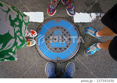 Sakata, Yamagata, Japan - Oct 5 2024, A close-up of a manhole cover with blue details and floral patterns, surrounded by the sneaker-clad feet of a family of four, at daytime, Sakata, Japan Sakata, Yamagata, Japan - Oct 5 2024, A close-up of a manhole cover with blue details and floral patterns, surrounded by the sneaker-clad feet of a family of four, at daytime, Sakata, Japan 137591030