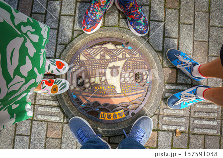 Sakata, Yamagata, Japan - Oct 5 2024, close-up view of a manhole cover with an colored image of ship sailing down river against of Sankyo Soko Storehouse, legs of family of four around, Sakata, Japan 137591038