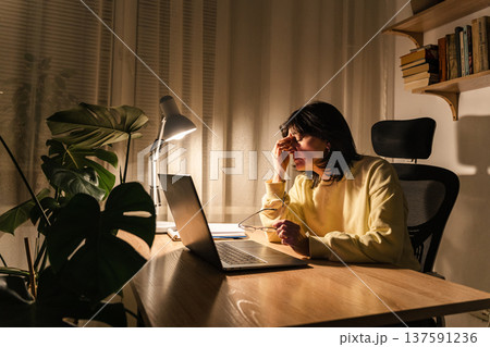 Exhausted woman experiencing burnout and stress while working on her laptop in a dark home office environment Exhausted woman experiencing burnout and stress while working on her laptop in a dark home office environment 137591236