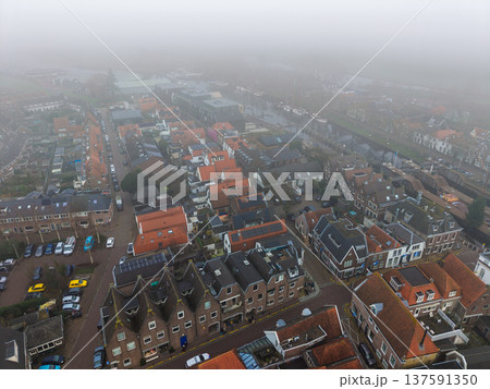 Atmospheric aerial view of a historic Dutch town shrouded in thick morning fog. Traditional brick houses with orange tiled roofs line a misty canal, creating a moody, picturesque European scene. Atmospheric aerial view of a historic Dutch town shrouded in thick morning fog. Traditional brick houses with orange tiled roofs line a misty canal, creating a moody, picturesque European scene. 137591350