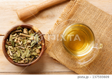A glass cup containing lemon balm tea, surrounded by dried leaves in a bowl and a wooden mortar on burlap fabric A glass cup containing lemon balm tea, surrounded by dried leaves in a bowl and a wooden mortar on burlap fabric 137593609