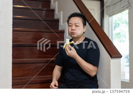 A man sits on the stairs at home, smiling while eating a banana as a healthy snack and source of natural energy after exercise. A man sits on the stairs at home, smiling while eating a banana as a healthy snack and source of natural energy after exercise. 137594759