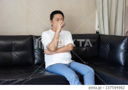 A man drinks a glass of water while sitting on a black leather sofa, representing daily hydration and a healthy lifestyle. 137594982