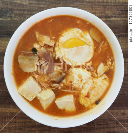 A bowl of spicy Korean tofu soup featuring soft tofu cubes, pork slices, kimchi, and a poached egg in rich red chili broth, a classic comforting dish known as Soondubu-jjigae at Asian restaurant A bowl of spicy Korean tofu soup featuring soft tofu cubes, pork slices, kimchi, and a poached egg in rich red chili broth, a classic comforting dish known as Soondubu-jjigae at Asian restaurant 137595805