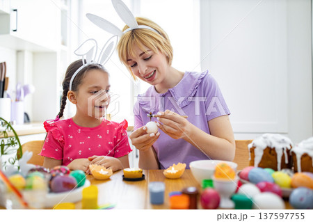 A mother and daughter wearing bunny ears happily decorate Easter eggs with wax and paints at a wooden table 137597755