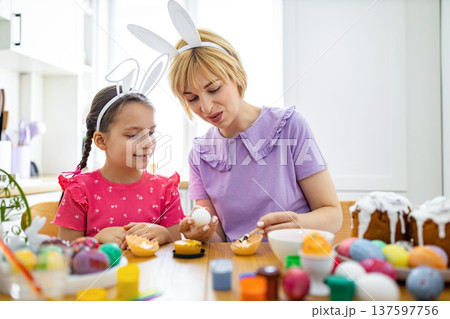 A mother and daughter wearing bunny ears are happily decorating Easter eggs with wax and paint at a wooden table A mother and daughter wearing bunny ears are happily decorating Easter eggs with wax and paint at a wooden table 137597756