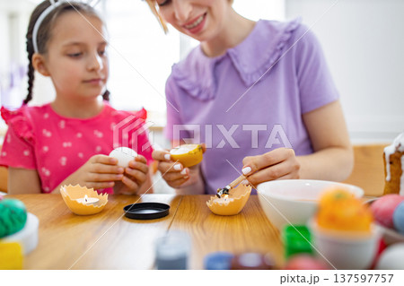 A mother and daughter are happily engaged in the traditional Easter activity of decorating eggs with wax and dyes at a wooden table A mother and daughter are happily engaged in the traditional Easter activity of decorating eggs with wax and dyes at a wooden table 137597757